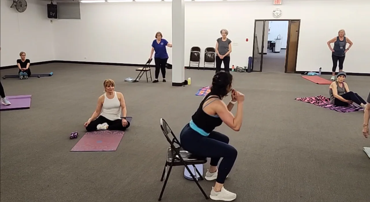 A group practicing chair-supported squats and floor exercises in a shared indoor space.